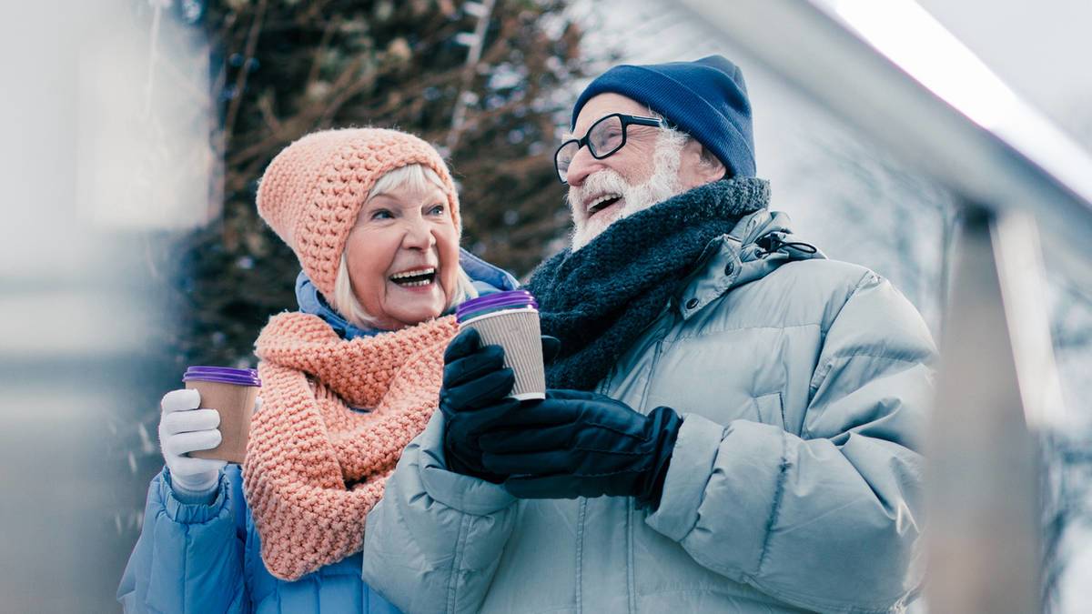 elderly couple in coats, hats, and gloves holding insulated cups and smiling at each other