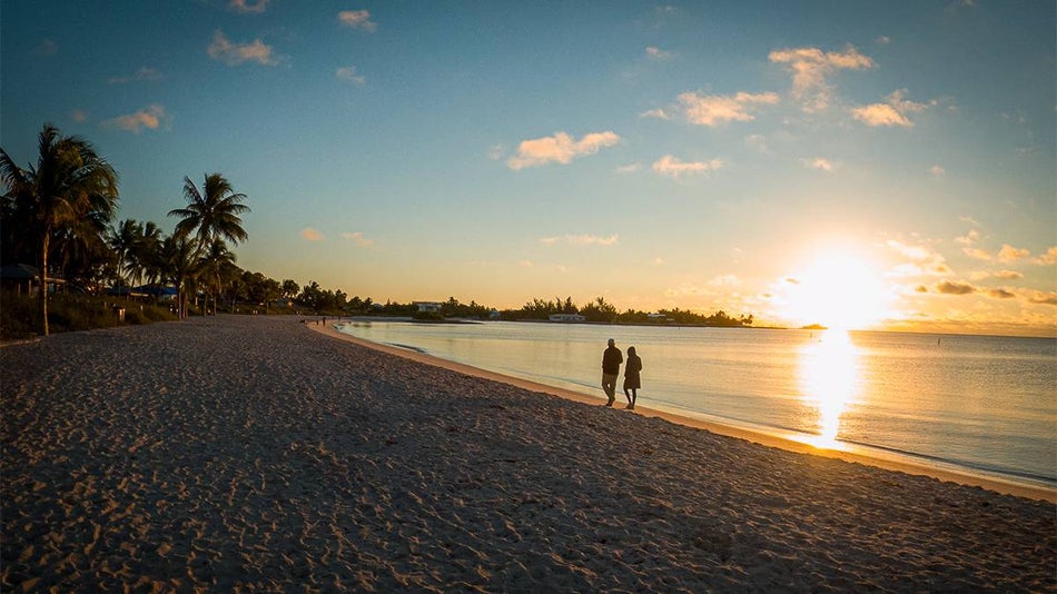 Wide shot of a couple walking where the water meets the sand at Sombrero Beach with a bright sunset in front of them in Key West, Florida, USA