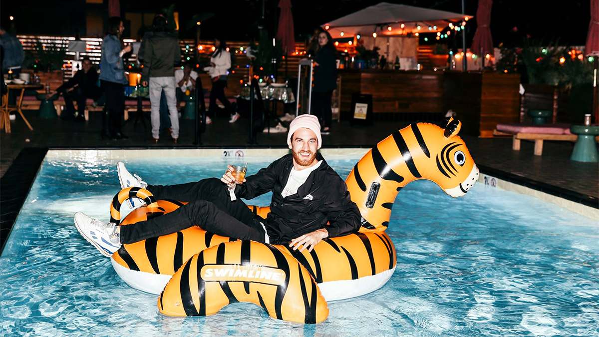 Adult man in a black outfit sitting on a giant inflatable tiger floaty in a small pool at night at the Broken Shaker in Los Angeles, California, USA