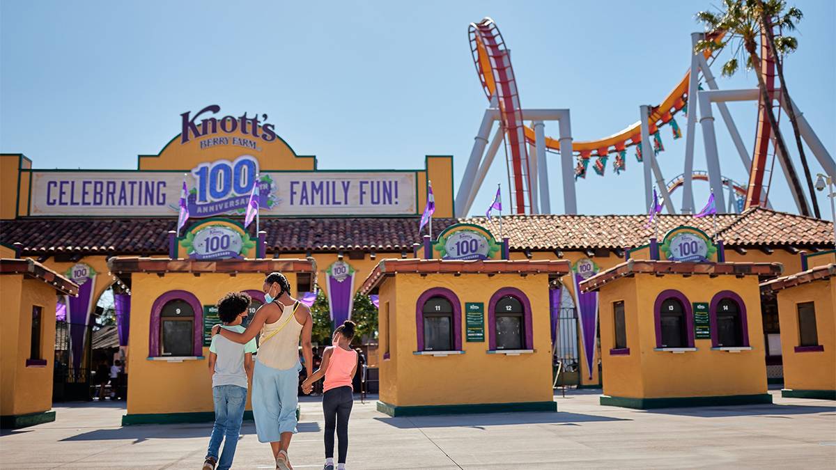 A mom and her two children walking into the yellow front gates of Knott's Berry Farm with a blue sky and roller coaster in the background in Los Angeles, California