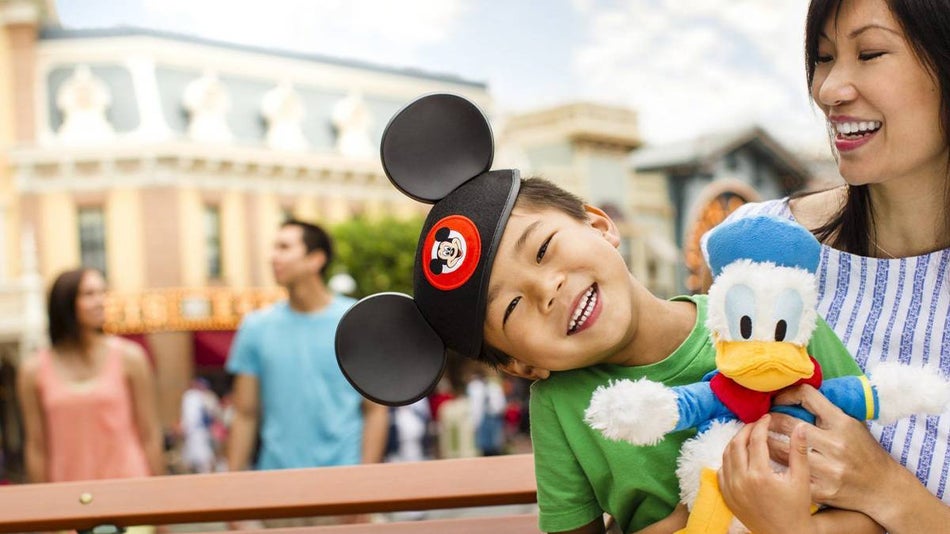Little boy with Mickey ears hat holding donald duck stuffed toy sitting on mother's lap in Disneyland in Los Angeles, California, USA