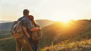 Man and woman hikers wearing backpacks hugging as the sun sets over hills