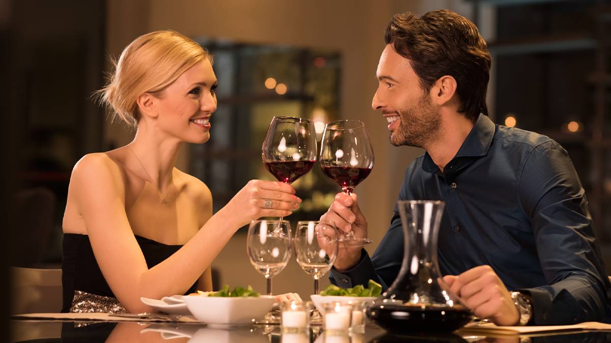 man and woman dressed nicely holding wine glasses sitting at dinner in front of twinkle lights