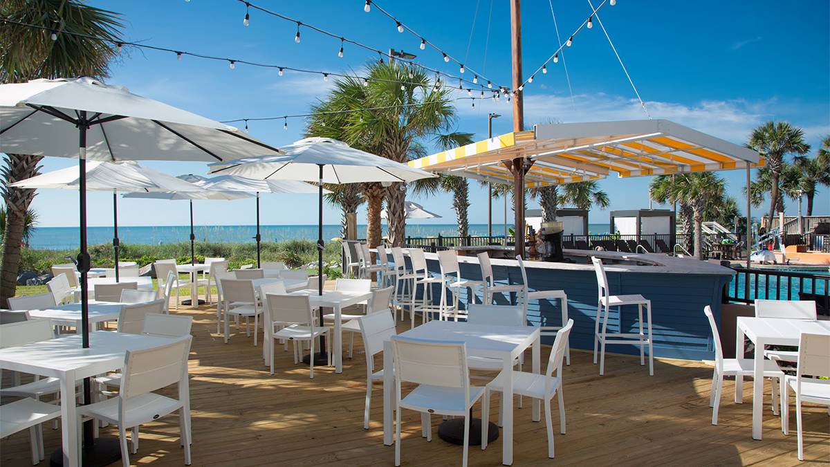 Wide shot of the outdoor dining area at Sea Captain's House, filled with white tables with white umbrella and a bar with the ocean and palm trees in the background in Myrtle Beach, South Carolina, USA
