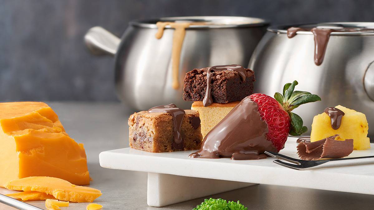 Close up photo of different fruits and deserts covered in chocolate fondu with fondu pots in the back ground and a large block of cheese to the right at The Melting Pot in Myrtle Beach, South Carolina, USA