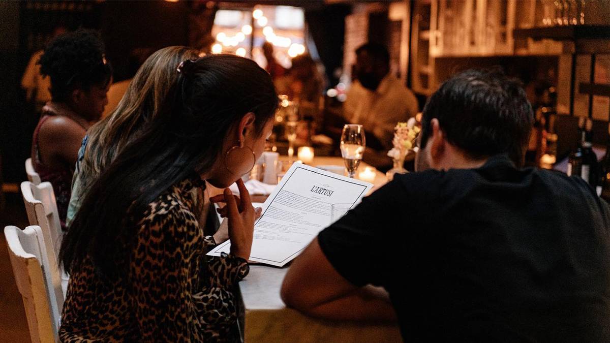 A couple looking over a paper menu with wine glasses infant of them in low light at L’Artusi in NYC, New York, USA