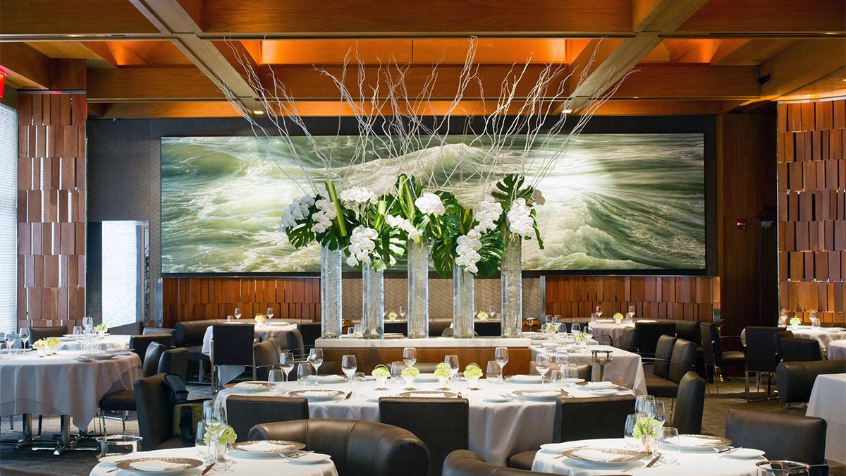 A dining room with a large floral center piece and tables surrounding it with a wide green mural on the back wall, each table is set with small flowers, gold plates, and wine glasses at Le Bernardin in NYC, New York, USA