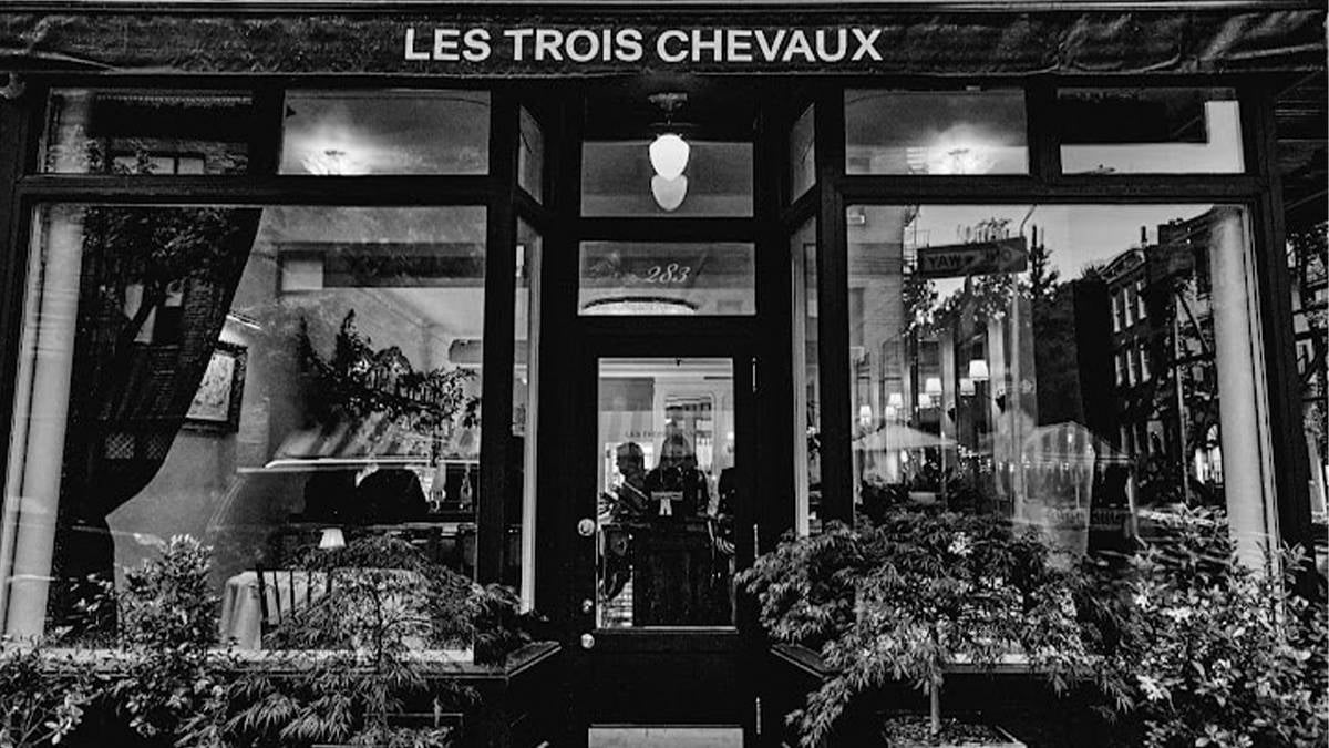 Exterior black and white view of Les Trois Chevaux with its large glass windows and the silhouettes of people inside in NYC, New York, USA