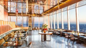 A large dining room with tables lining the left and right side and large tall glass windows and a reflective ceiling, each table is set and has flowers and the sun is setting outside, the room has an orange glow at Peak in NYC, New York, USA