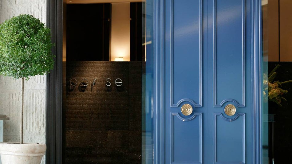 An entrance with two large blue doors with windows on either side and a small tree to the right with a sign reading Per Se visible through the window as the entrance to Per Se in NYC, New York, USA