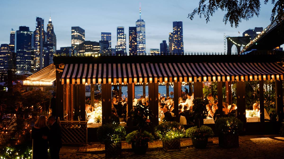 Wide shot of the exterior of The River Cafe at dusk with their lights on and a dining room full of people and the city skyline behind it in NYC, New York, USA