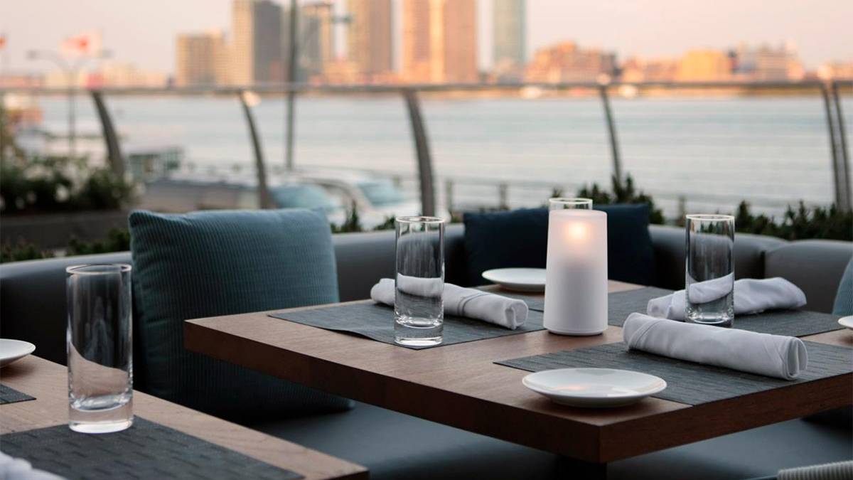 Close up photo of a square wooden tables with a white candle in the middle with 3 place settings at sunset with the river and city in the background at Riverpark in NYC, New York, USA