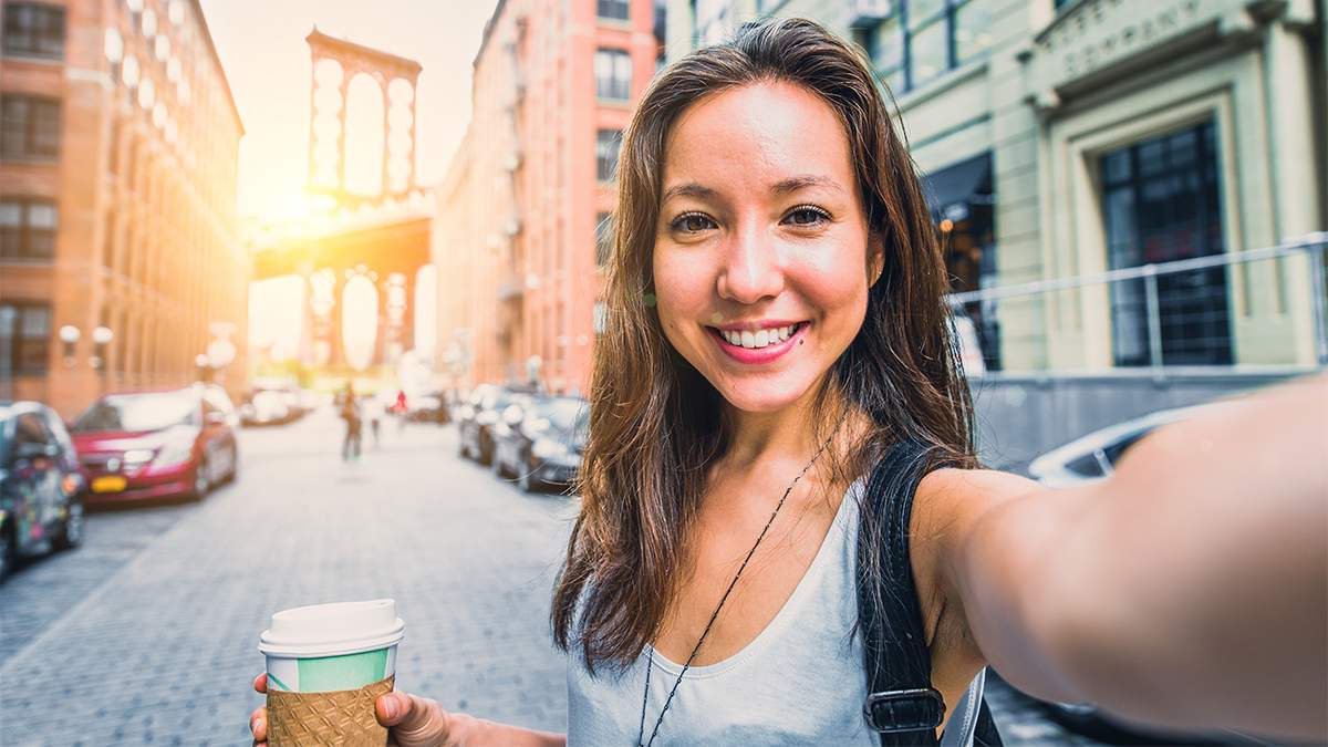 woman holding coffee and standing on street with bridge and sunset in background in NYC, New York, USA