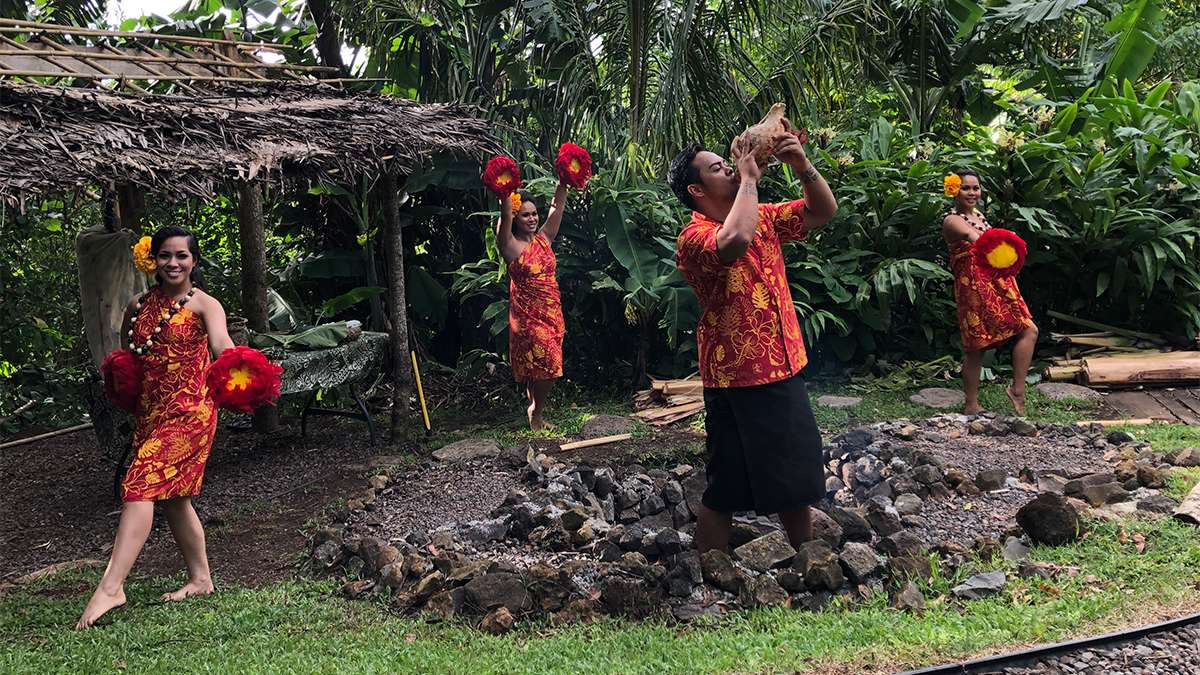 Three women in orange and red hula costumes and a man in a matching shirt playing a conch shell surrounded by greenery at the Experience Nutridge Luau on Oahu, Hawaii, USA