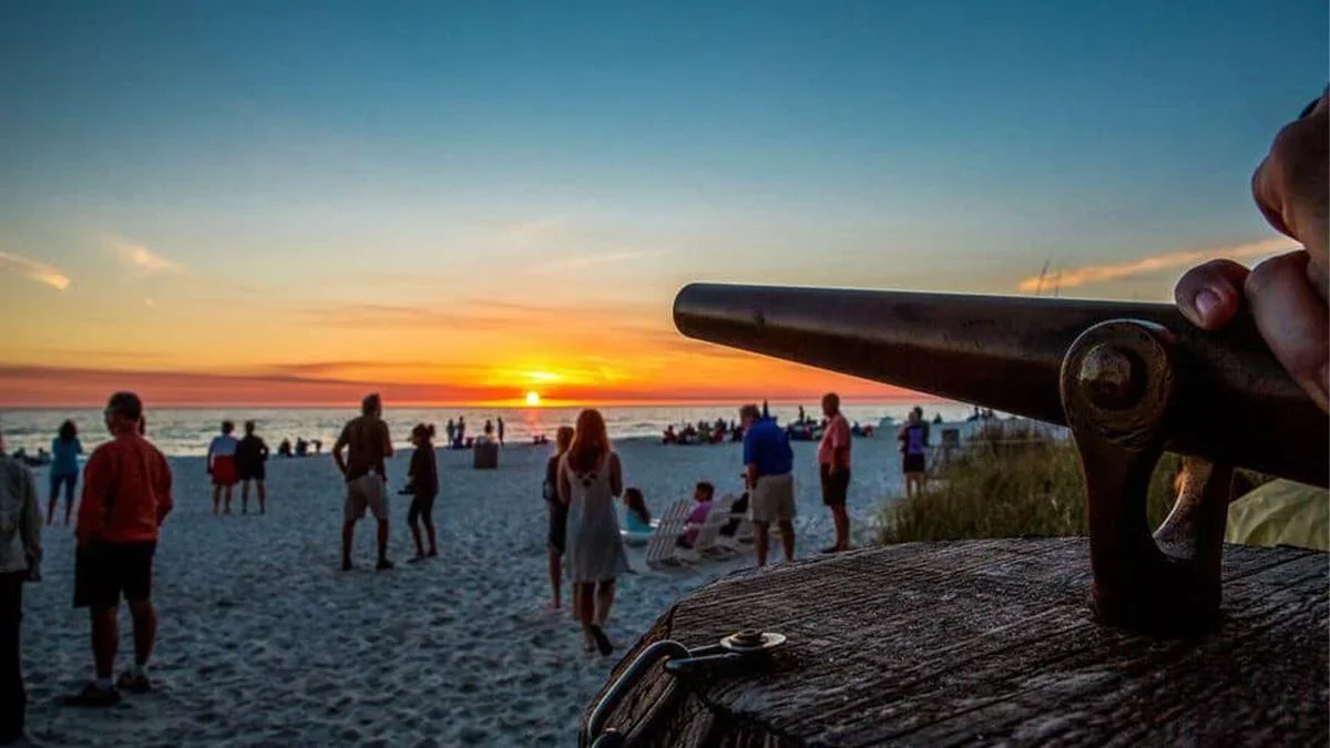 Close up of the cannon with people on the beach and a bright orange sunset in the background at Sunset Celebration Cannon at Schooners Beach Club in Panama City, Florida, USA
