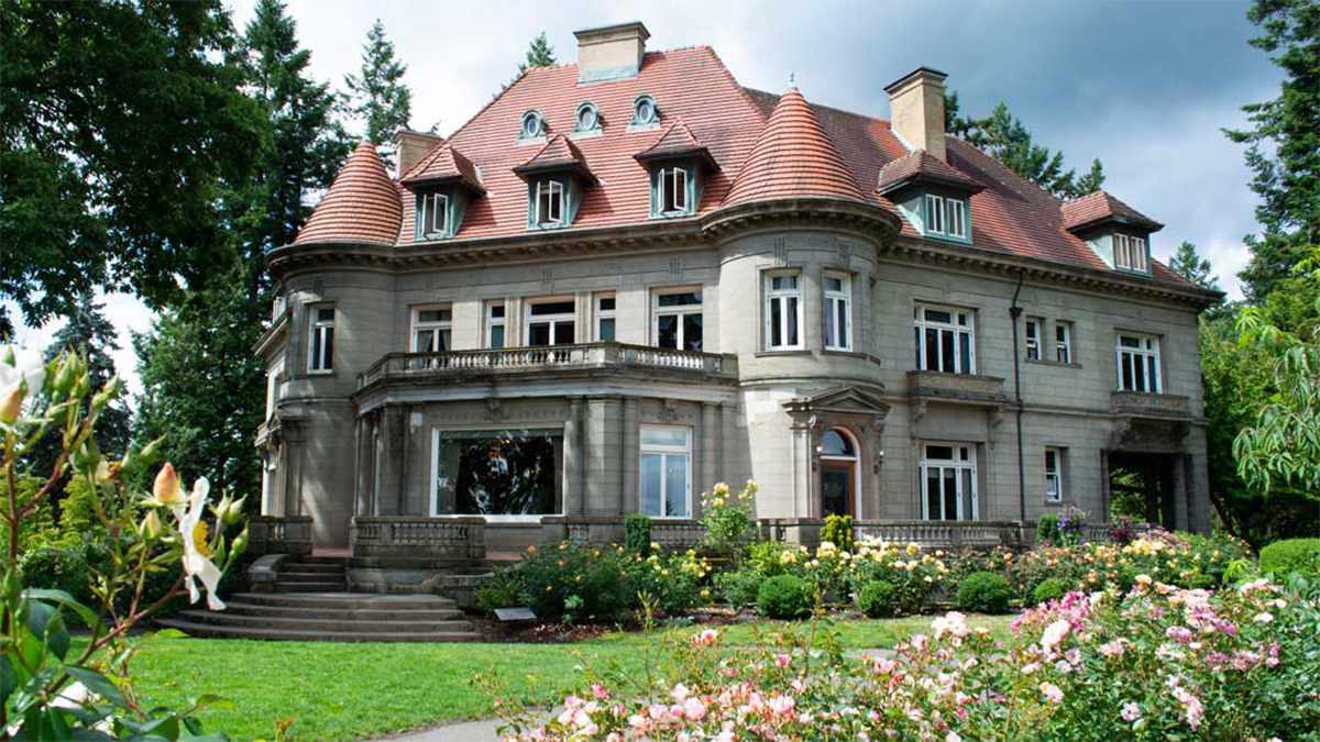 Wide shot of the stone exterior of Pittock Mansion with a burnt red roof on a sunny spring day surrounded by different types of bushes and flowers in Portland, Oregon, USA