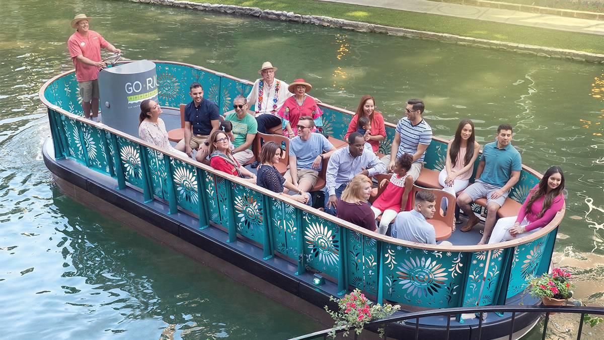 A river boat full of people on a tour with a guide standing at the back of the boat on a sunny day in San Antonio, Texas