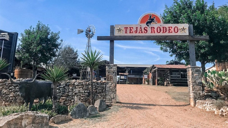 Entrance to Tejas Rodeo with their sign over a walk way and a bull statue on the left with trees mixed in and building in the background