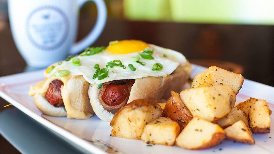 Close up of two bacon wrapped hotdogs in buns with a runny eggs on top and breakfast potatoes from Breakfast Republic in San Diego, California, USA