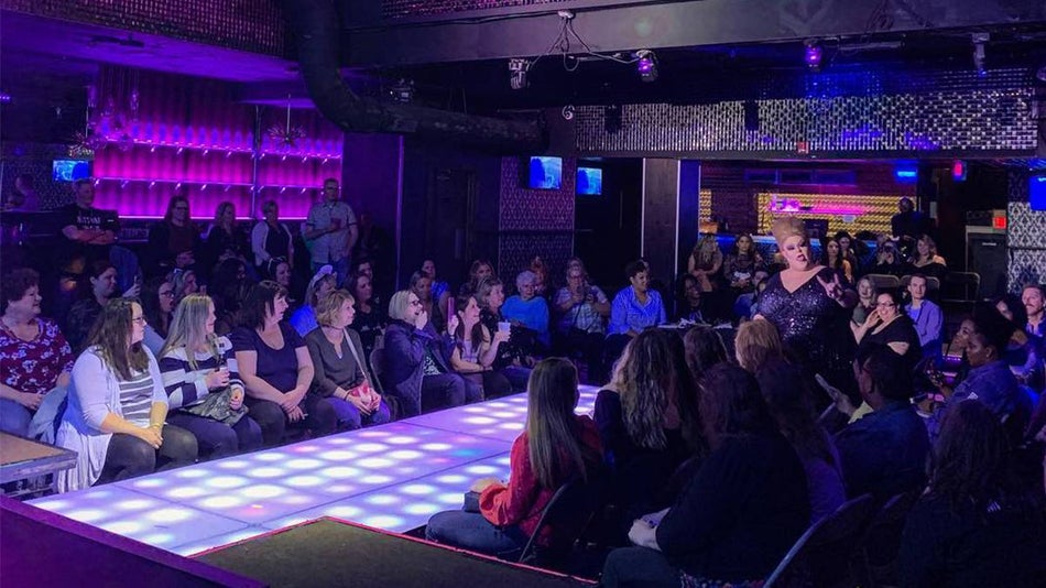 View of a catwalk with women on either side of it with low light and a drag queen dressed in black talking to the crowd at Hunk-O-Mania in San Diego, California, USA