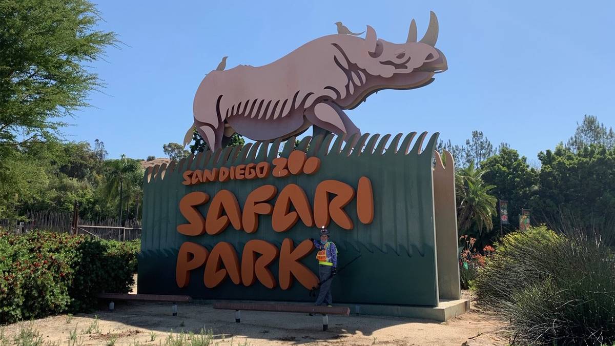 Zoo employees standing with a large green sign with orange lettering saying "San Diego Zoo Safari Park" with a rhino on top of it on a sunny day in San Diego, California