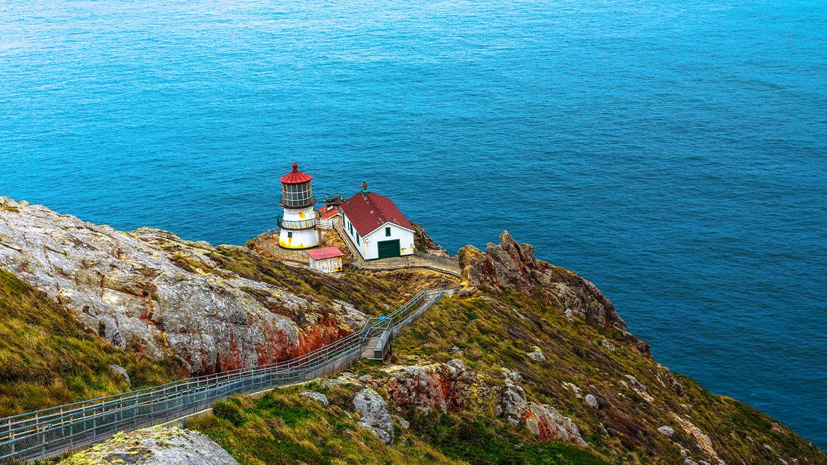 View looking down on a building and light house with a red roof at Point Reyes Station with a bright blue ocean behind it in San Francisco, California, USA