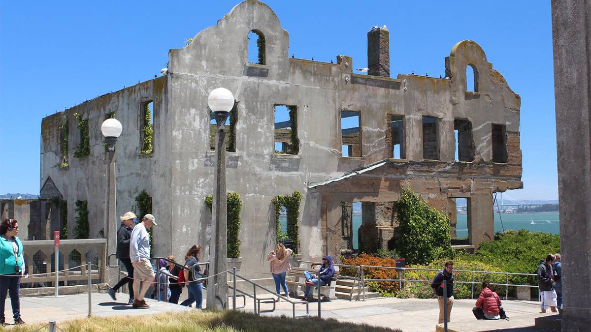 Ground view of ruins of the old Warden House with green shrubs and people walking in front of it on a sunny day on Alcatraz Island in San Francisco, California, USA