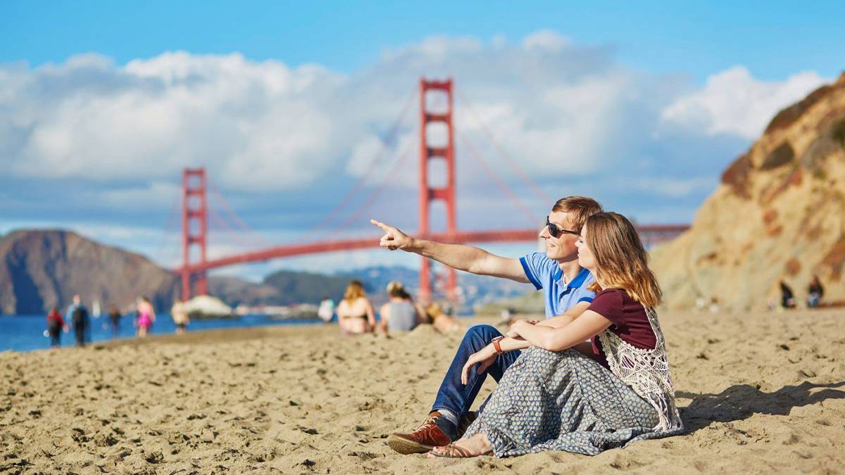 Couple sitting together on beach during daytime pointing at sky with Golden Gate Bridge in background in San Francisco, California, USA