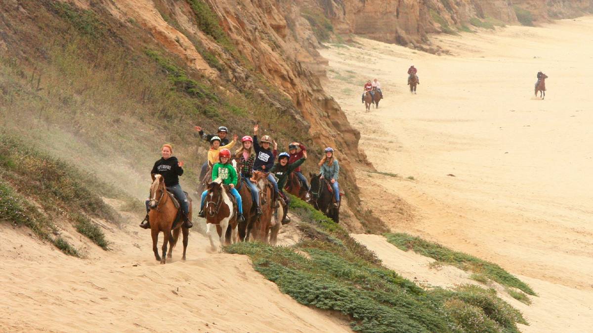 Wide shot of a group of people riding horses in that sand at Sea Horse Ranch, Half Moon Bay in San Francisco, California, USA