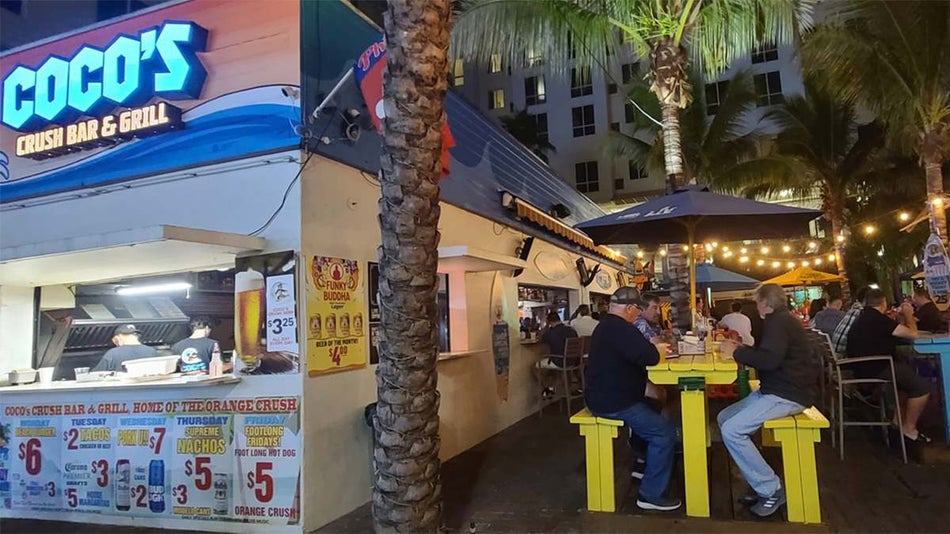 View of the side of Coco’s Crush Bar & Grill with their outdoor dining area to the right with yellow and blue tables at night in Tampa, Florida, USA