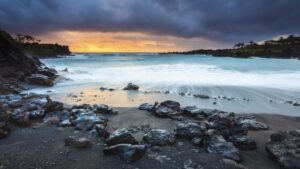 Black sand beach with rocks and waves coming in under a cloudy grey sky with orange sunset in the distance at Waianapanapa state park maui