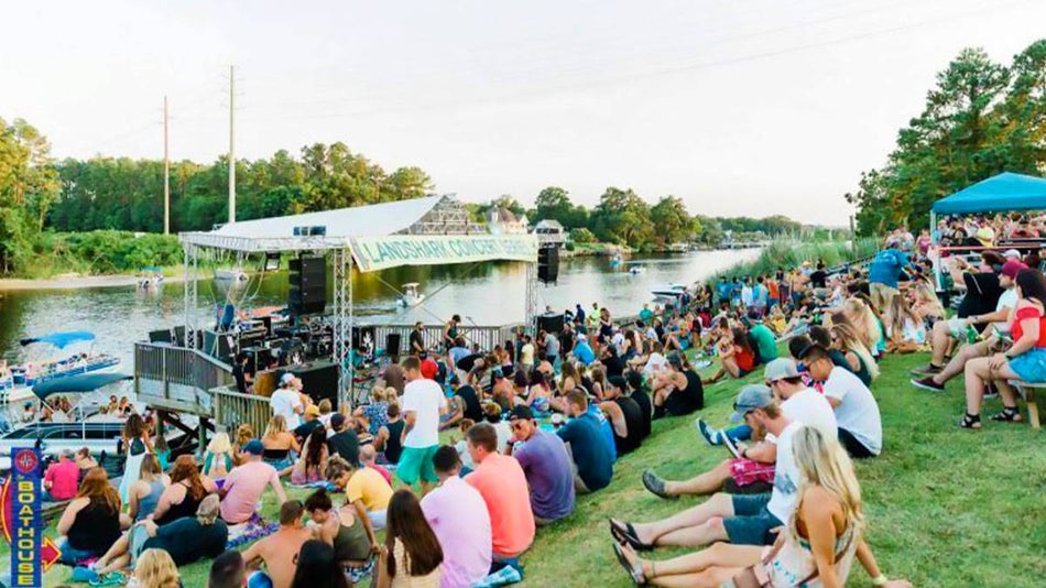 crowd sitting on grass in front of stage positioned at edge of shore with water in background and view of trees and greenery on other side