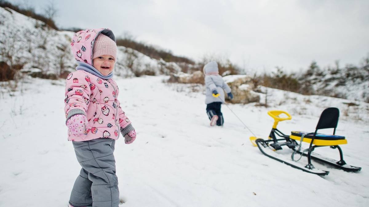 little girl in a pink and grey snowsuit smiling at the camera with a yellow and black sled in the background being pulled by small child in grey and black snowsuit in Alaska