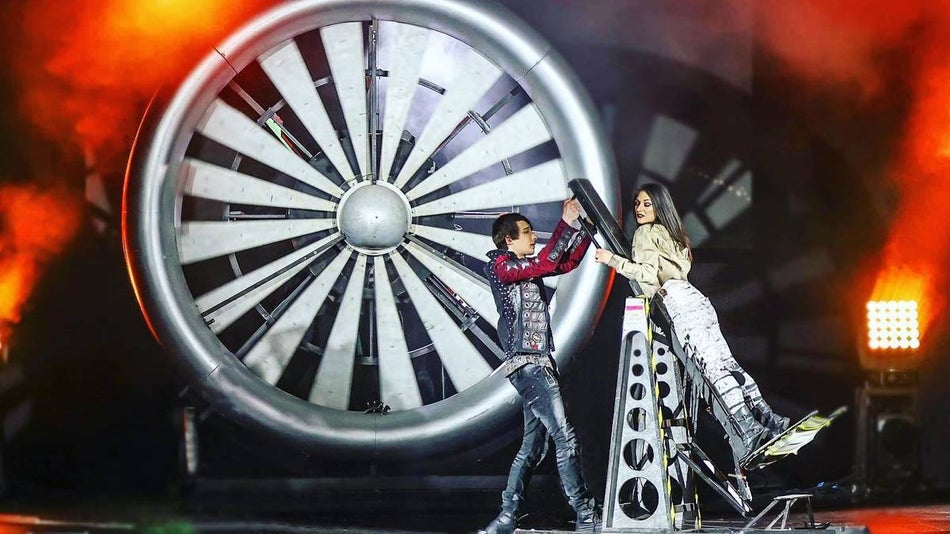 Reza with stage assistant performing a magic trick with a jet engine in the background and red colored stage smoke swirling around them