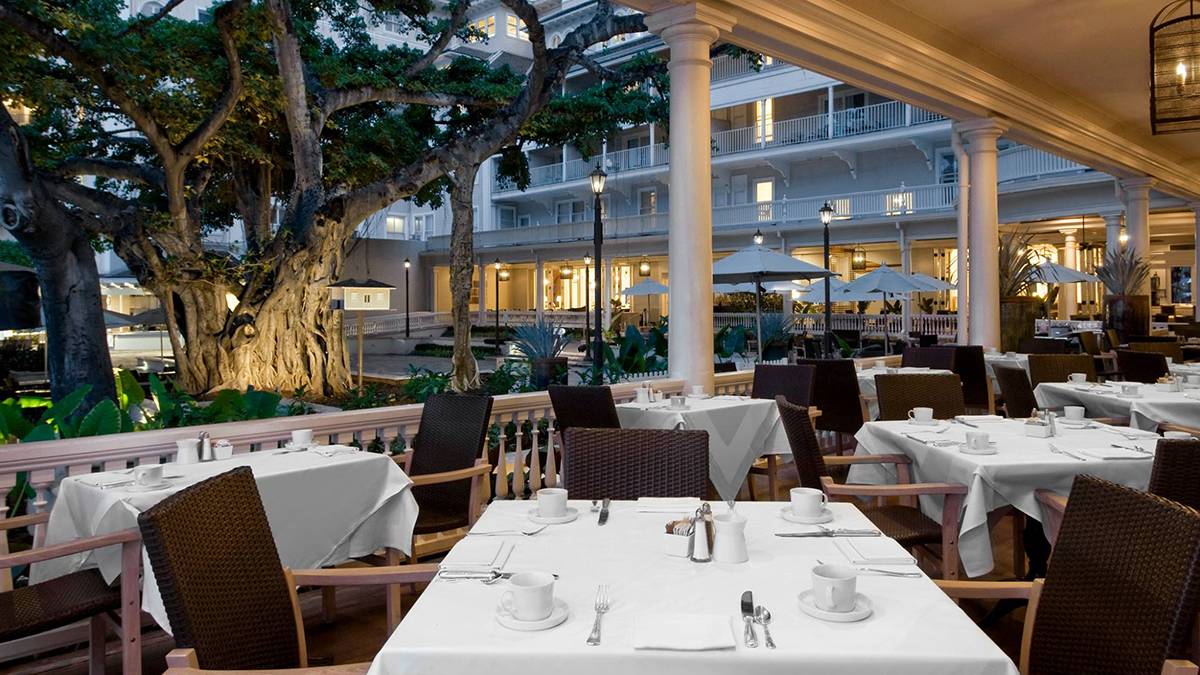 Outdoor dining with lots of tables covered in white table cloths and dark drown chairs with a huge tree on the other side of a white fence behind the tables at dusk at Beachhouse at the Moana in Honolulu, Hawaii, USA