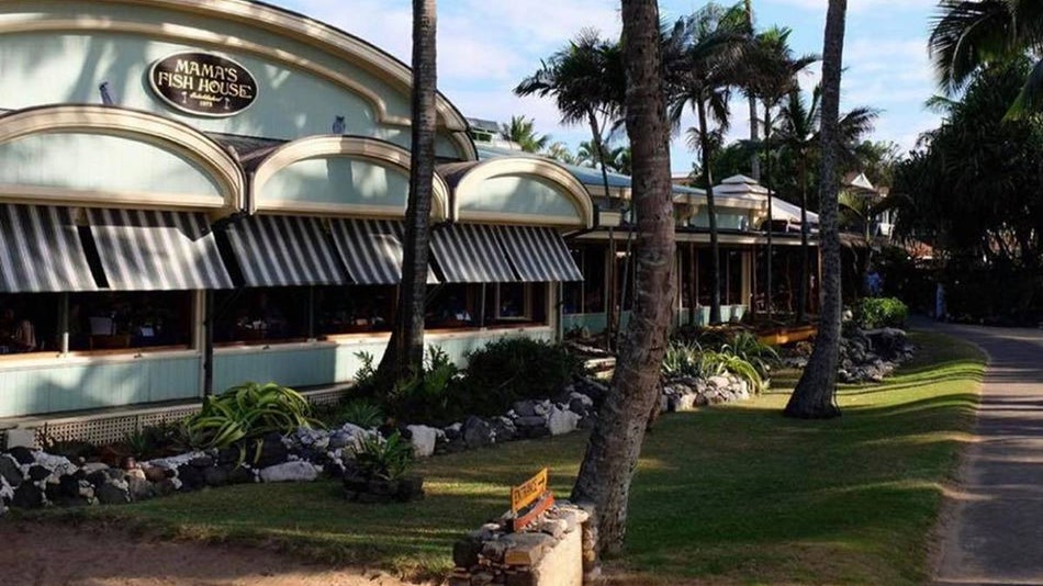 Wide exterior shot of Mama’s Fish House, painted a light blue green color and landscaped with lots of tropicals plants and palm trees in Honolulu, Hawaii, USA