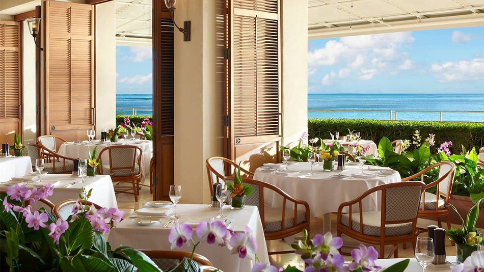 A dining room with white cloth covered tables and wooden chairs decorated with colorful flowers and large openings in the wall and an ocean view at Orchids in Honolulu, Hawaii, USA