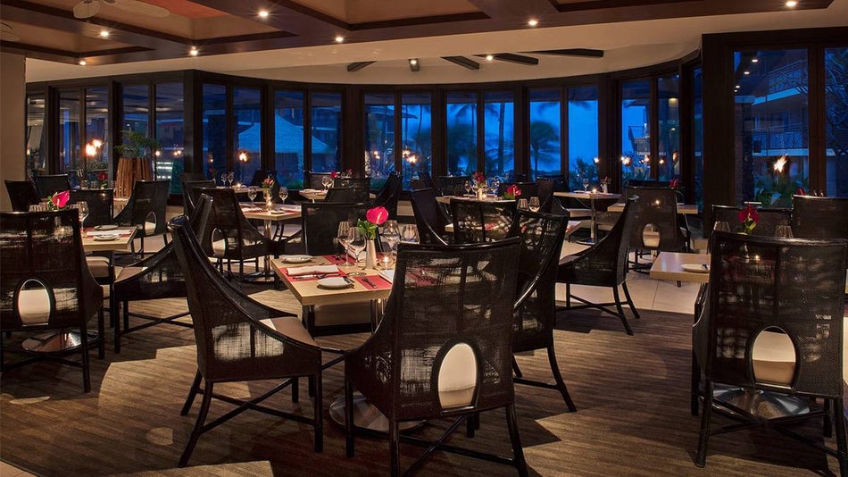A large dining rom filled with dark chairs and lighter wooden tables with flowers on them and large window in the back showing nightfall at Red Salt in Kauai, Hawaii, USA