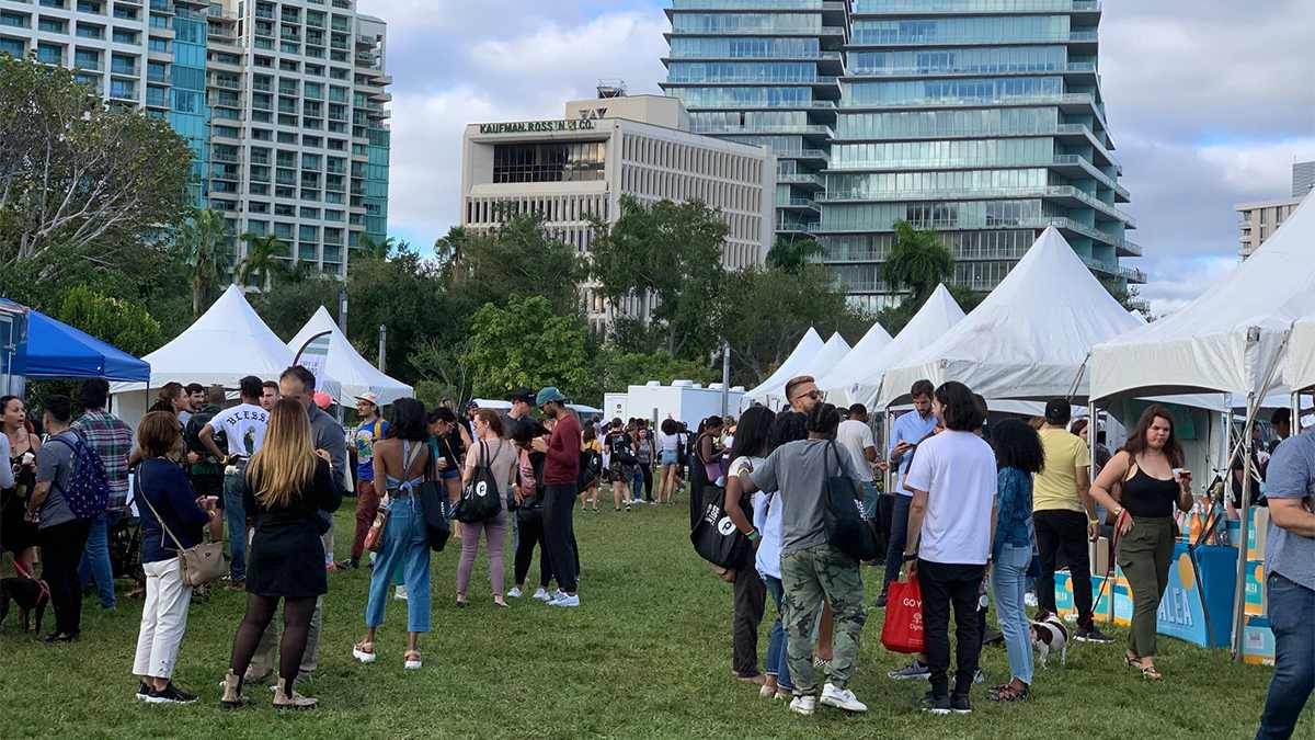 Wide shot of lots of people drinking and eating and white tents lining the left and right sides at the Coconut Grove Food and Wine Festival with large buildings in the background in Miami, Florida, USA