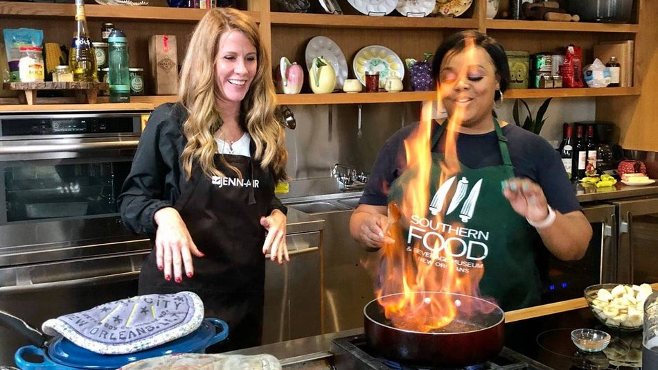 Two women in aprons cooking, one with a pan that has flames in it, at the Southern Food and Beverage Museum in New Orleans, Louisiana