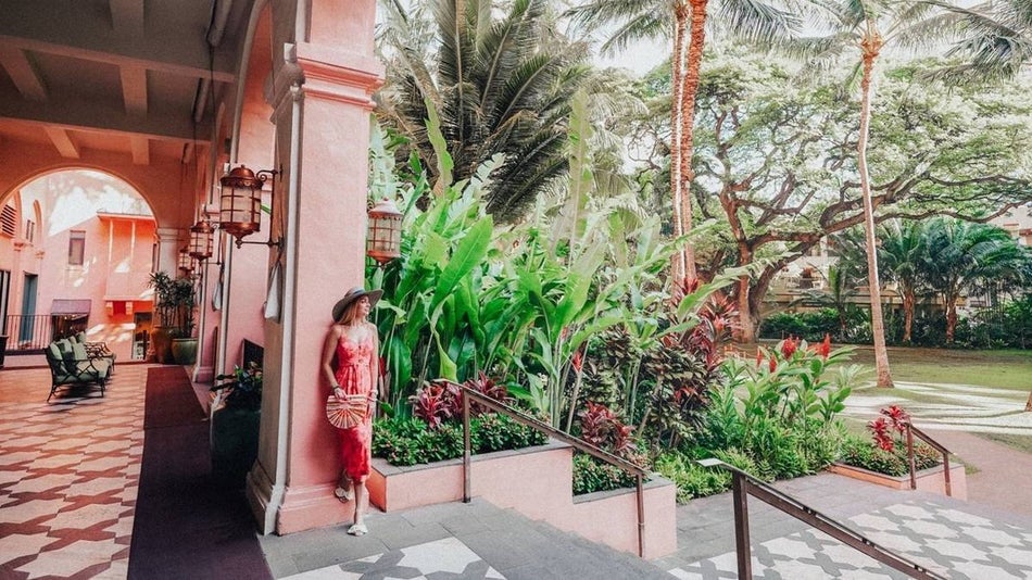 Woman leaning against a pink wall surrounded by colorful hawaiian foliage at the 'pink palace' Royal Hawaiian in Oahu.