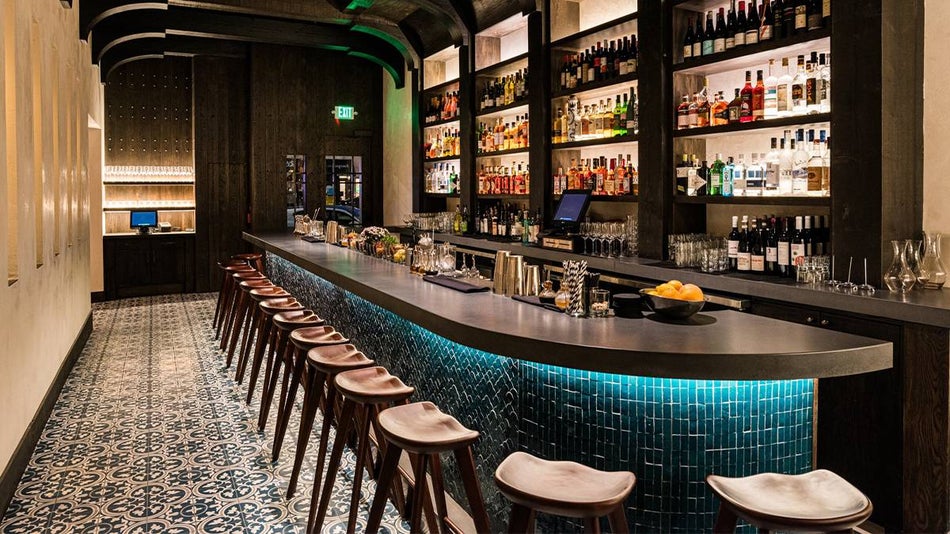 A long bar with blue tile, a black countertop, and wooden stools with different fruits and cups lining the bar and a wall full of alcoholic behind the bar at Aziza in San Francisco, California, USA
