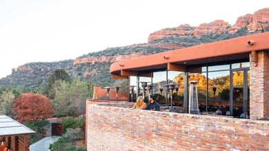 A wide shot of a couple sitting in the outdoor dining area of Che Ah Chi with large red rock formations behind the brick building in Sedona, Arizona, USA