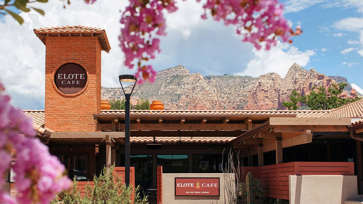The front of the Elote Cafe, beige walls, red and brick accents with a Rocky Mountain behind it and pink flowers close to the camera in Sedona, Arizona, USA