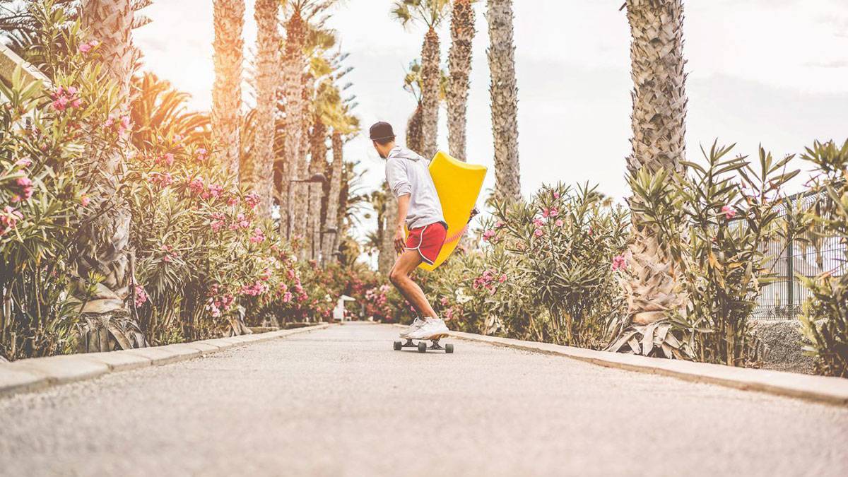 man skateboading down sidewalk with surf board and palm trees