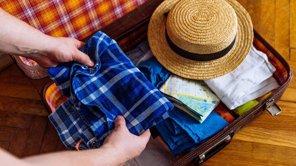 Man setting a blue checked shirt into a suitcase that has a hat and assortment of other clothes and belongings