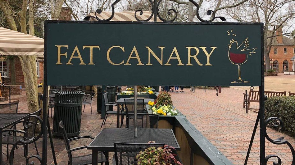 Close up of a green and gold sign that says Fat Canary with a wine glass that morphs into a bird on it with their outdoor seating in the background in Williamsburg, Virginia, USA