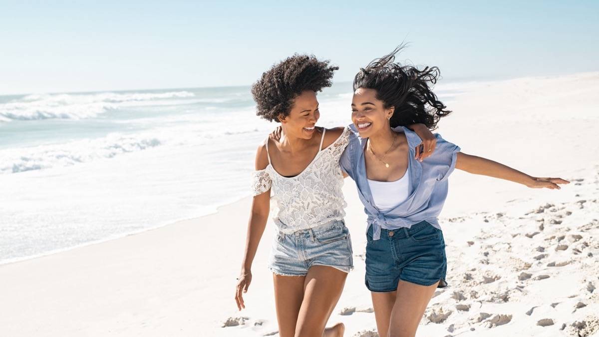 Two women with their arms around each other walking down the beach with the ocean in the background