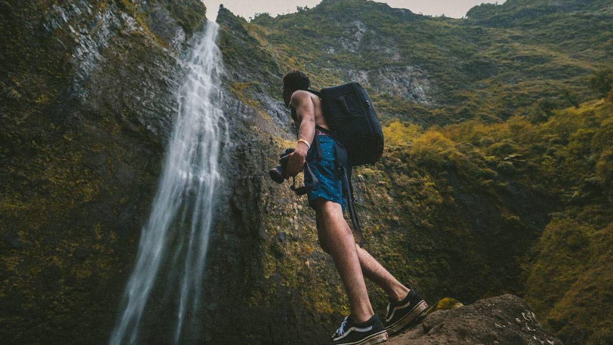 man standing under Hanakapiai Falls with hiking backpack and camera in Kauai, Hawaii, USA