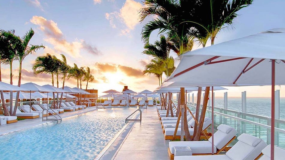 View of the pool and lounge chairs around it with palm trees and the ocean in the background at sunset at 1 Hotel South Beach Rooftop in Miami, Florida, USA